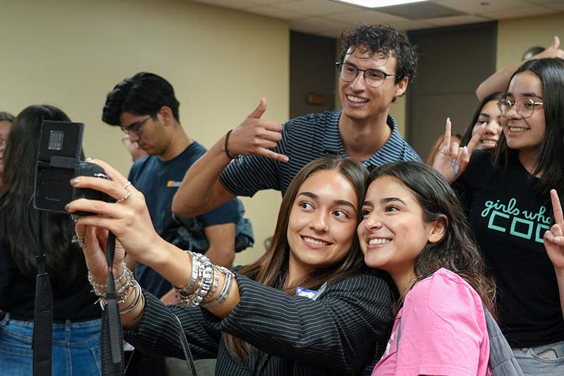 Five attendees pose together in front of a decorative balloon backdrop during a UTEP Engineering homecoming event, making hand gesture signs while wearing College of Engineering apparel and standing for a group photo.